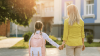 Mutter und Kind mit Schulranzen stehen Hand in Hand vor einer Schule