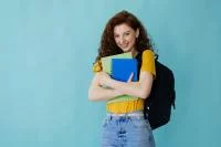 Eine Abiturientin in einem gelben Shirt hält Bücher in der Hand und steht vor einer türkisen Wand